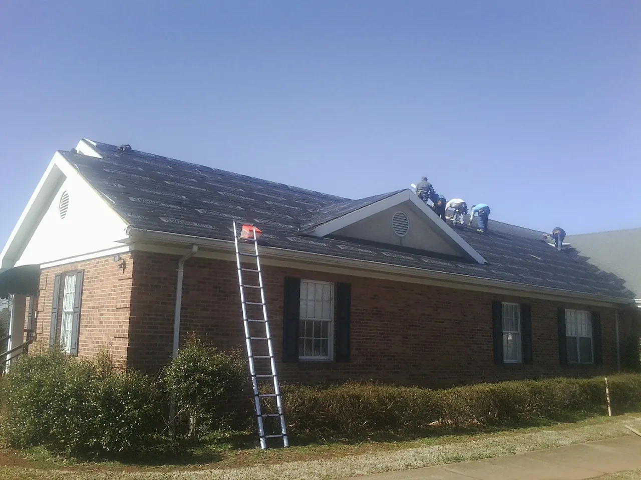 Crew tearing shingles off a brick ranch roof with a ladder set — Emergency Roof Repair work by our crew in Oakham