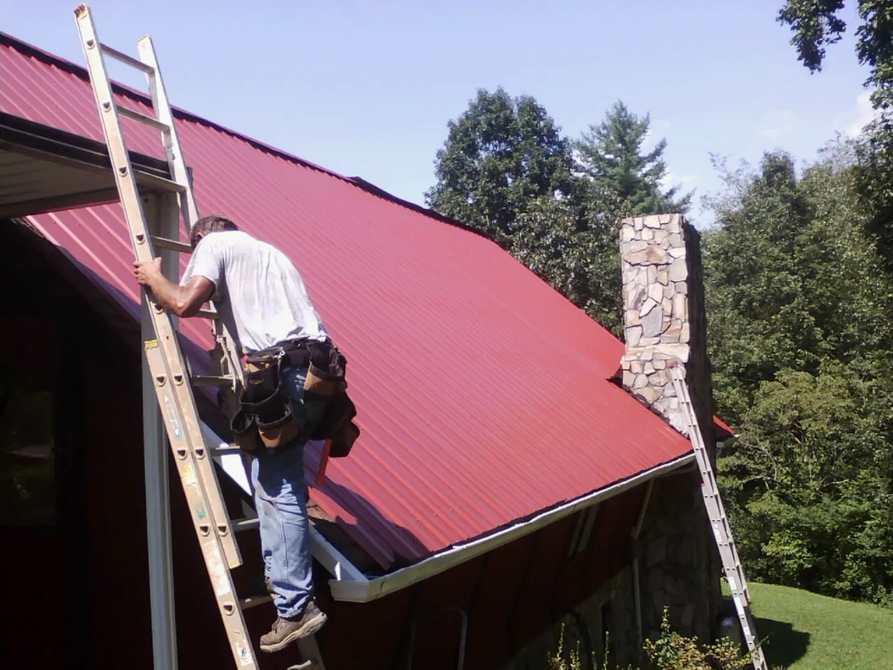 Roofer on a ladder beside a red metal roof and stone chimney — Soffit & Fascia Repair by Roofing Brothers in Oakham